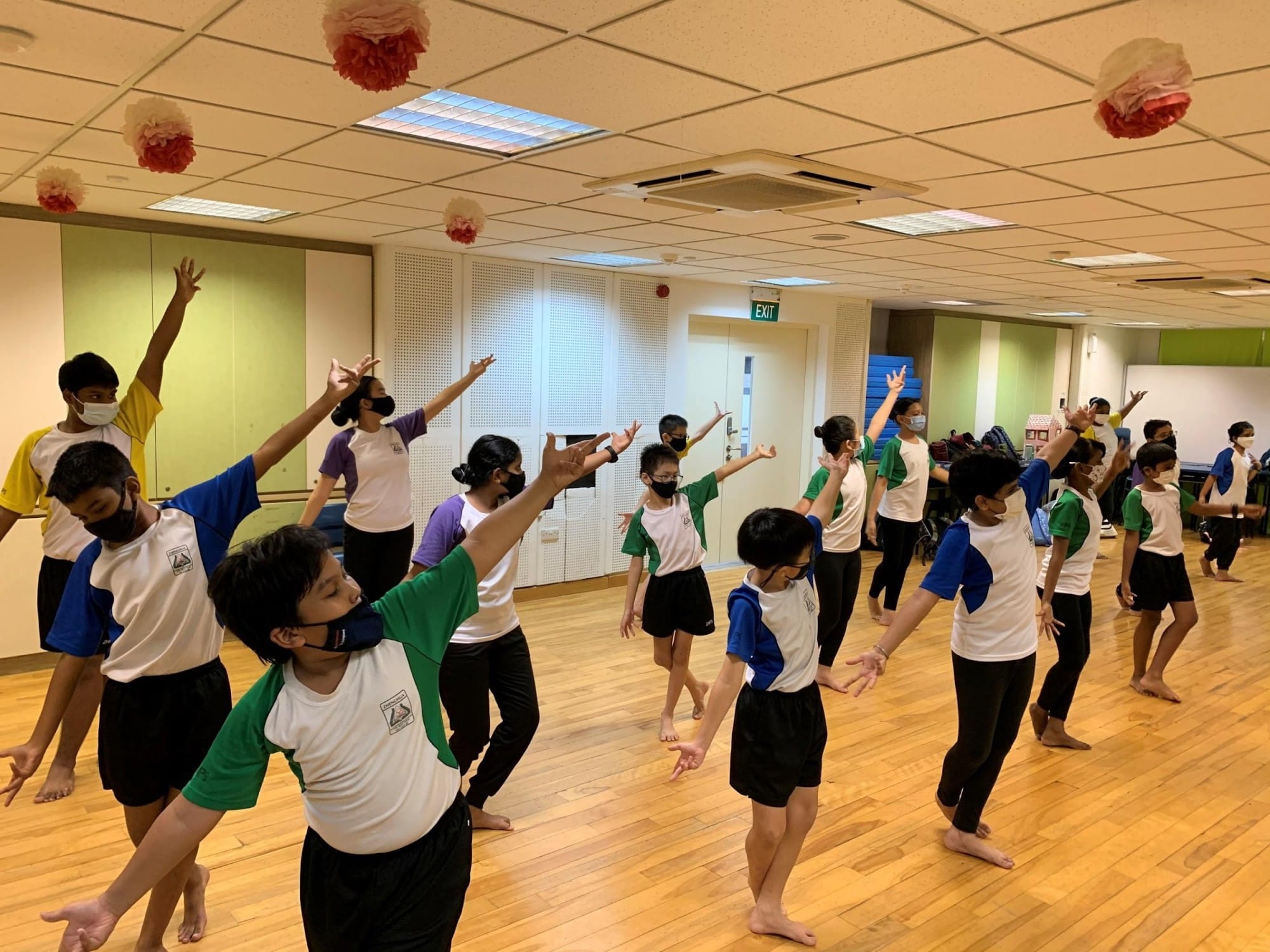 Children wearing masks practice dance moves barefoot in an indoor studio with paper pompoms on the ceiling.